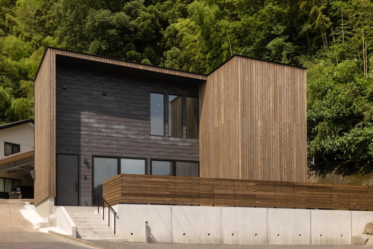 豊かな緑を背に黒い外装材と木目のパネルを組み合わせたコントラストが美しい二階建て