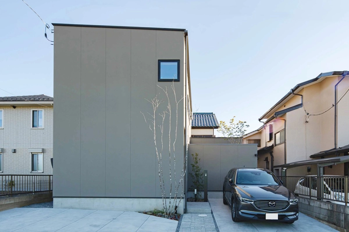 窓を最小限に抑えたグレーのミニマルな外壁と並列駐車された乗用車のある玄関アプローチ