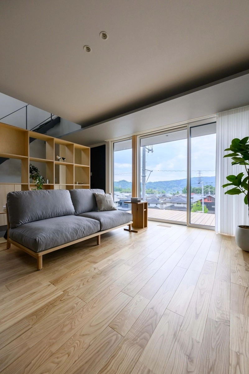 明るい無垢材の床にグレーのソファを配した高台の景色を望む開放的なリビング