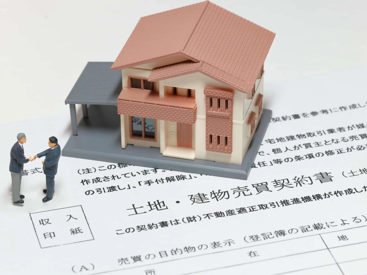 土地建物売買契約書の上に置かれた住宅模型と握手する男性2人のミニチュア