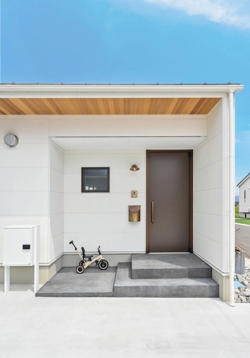白い外壁に木目の軒天井が映える玄関ポーチと、三輪車が置かれたコンクリートの土間