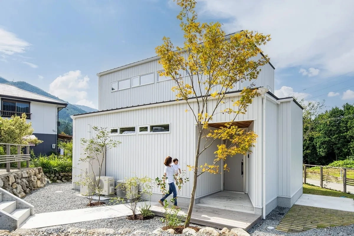 紅葉した木のあるアプローチを歩く親子と白い外壁の住宅