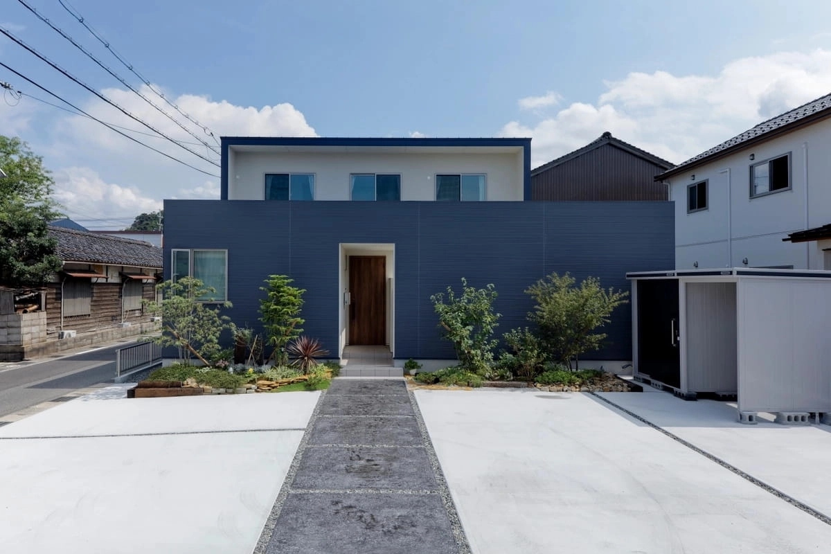 コンクリートの駐車場と植栽豊かなアプローチのある青い住宅